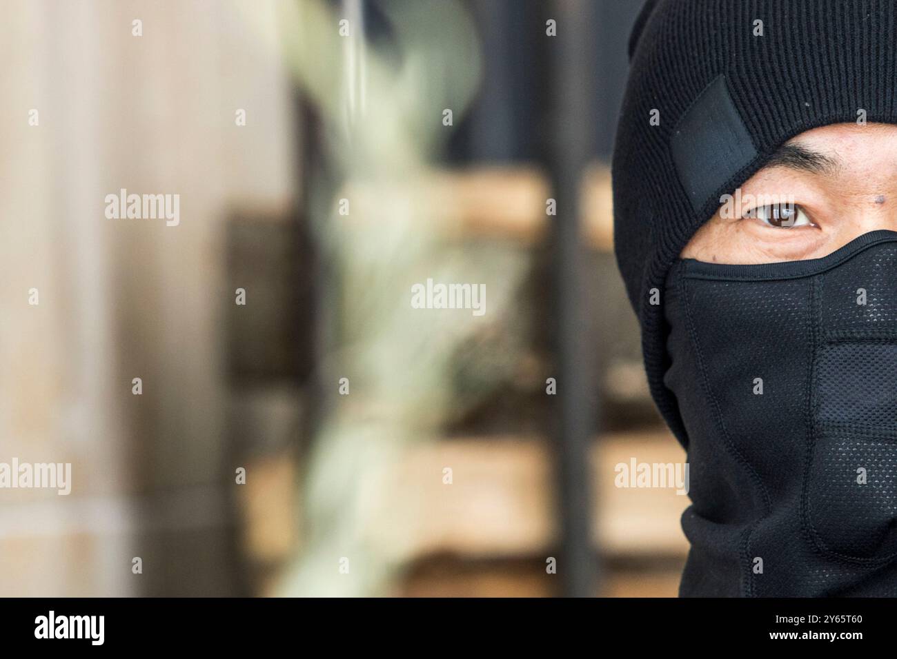 Primo piano di un misterioso uomo asiatico in balaclava nera, con un occhio visibile, che simboleggia il segreto o la protezione. Dietro, lo sfondo sfocato suggerisce t Foto Stock