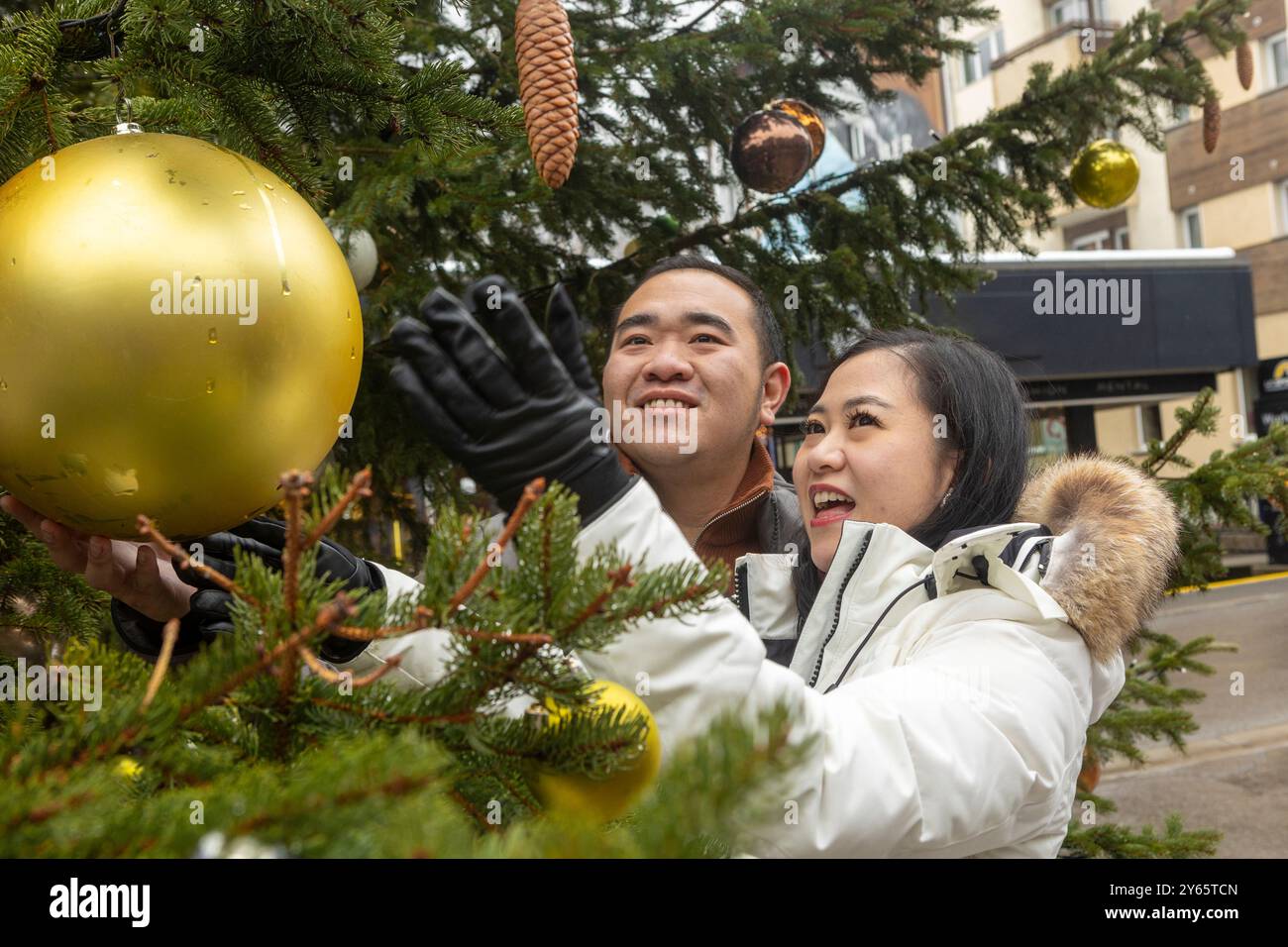 Allegra coppia asiatica decora un lussureggiante albero di Natale a Zermatt, aggiungendo un grande ornamento dorato mentre vi godete momenti festivi insieme in una pittoresca u Foto Stock