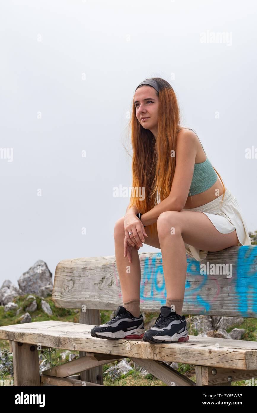 Una giovane donna siede su una panchina di legno nelle Asturie, guardando lontano con attenzione con montagne nebbiose sullo sfondo. Foto Stock