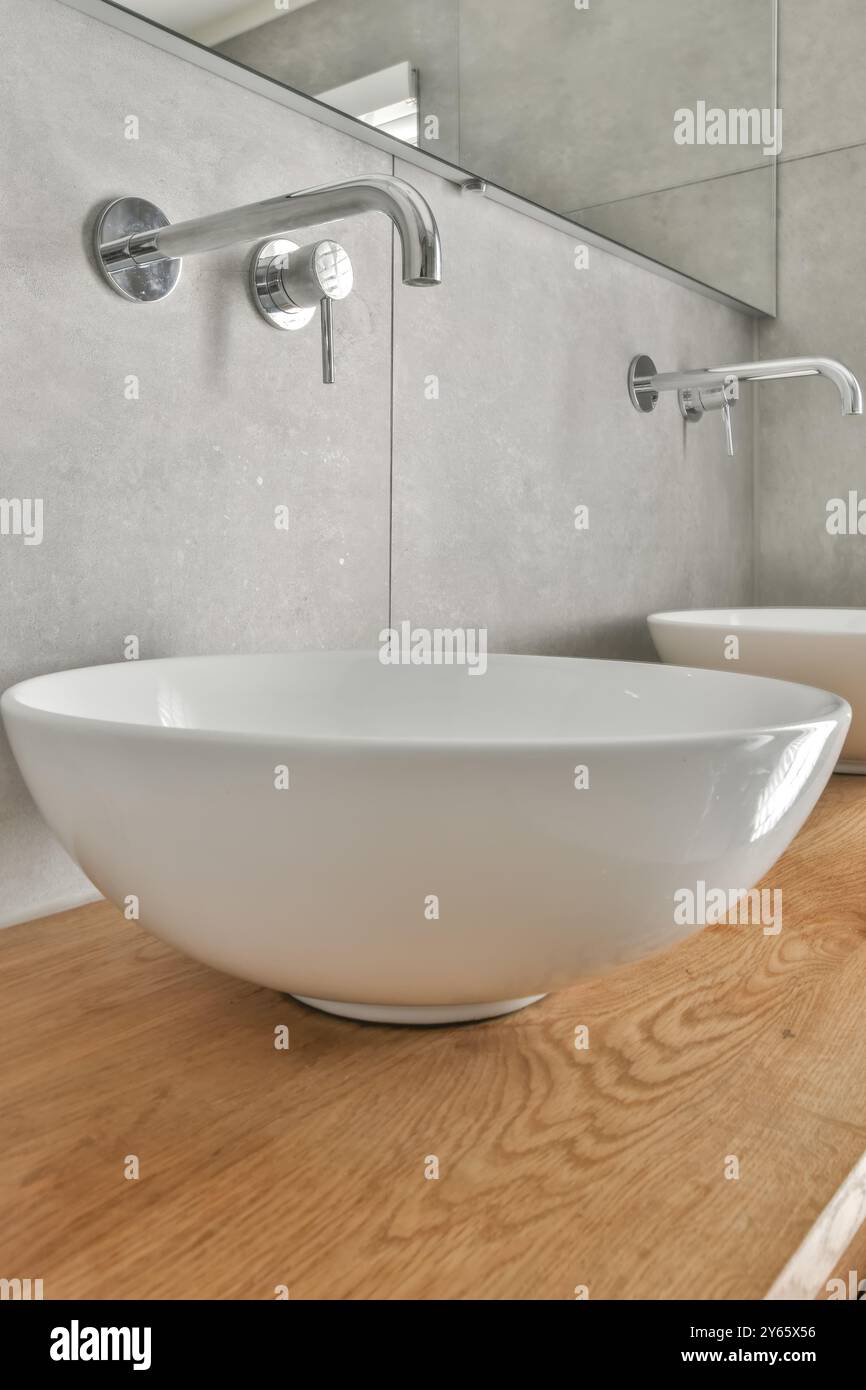 Un bagno elegante e moderno con un lavandino rotondo in cima ad un lavandino in legno, completato da rubinetti cromati montati a parete e arredi minimalisti. Foto Stock