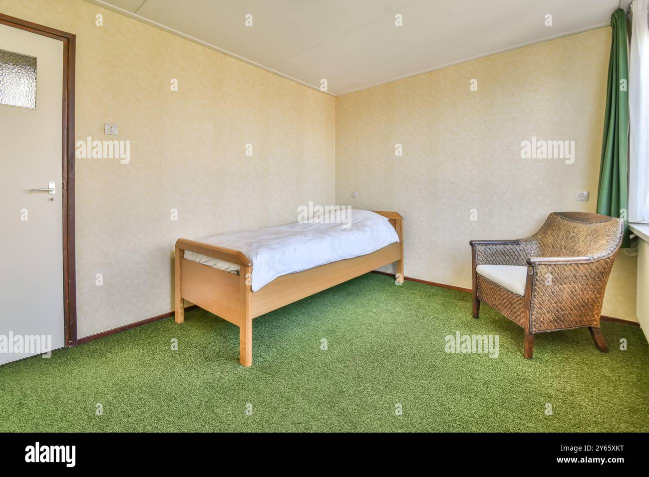 Una camera da letto semplice presenta un letto singolo in legno con biancheria bianca e una poltrona in vimini, con tappeti verdi e pareti a tappeto. Foto Stock