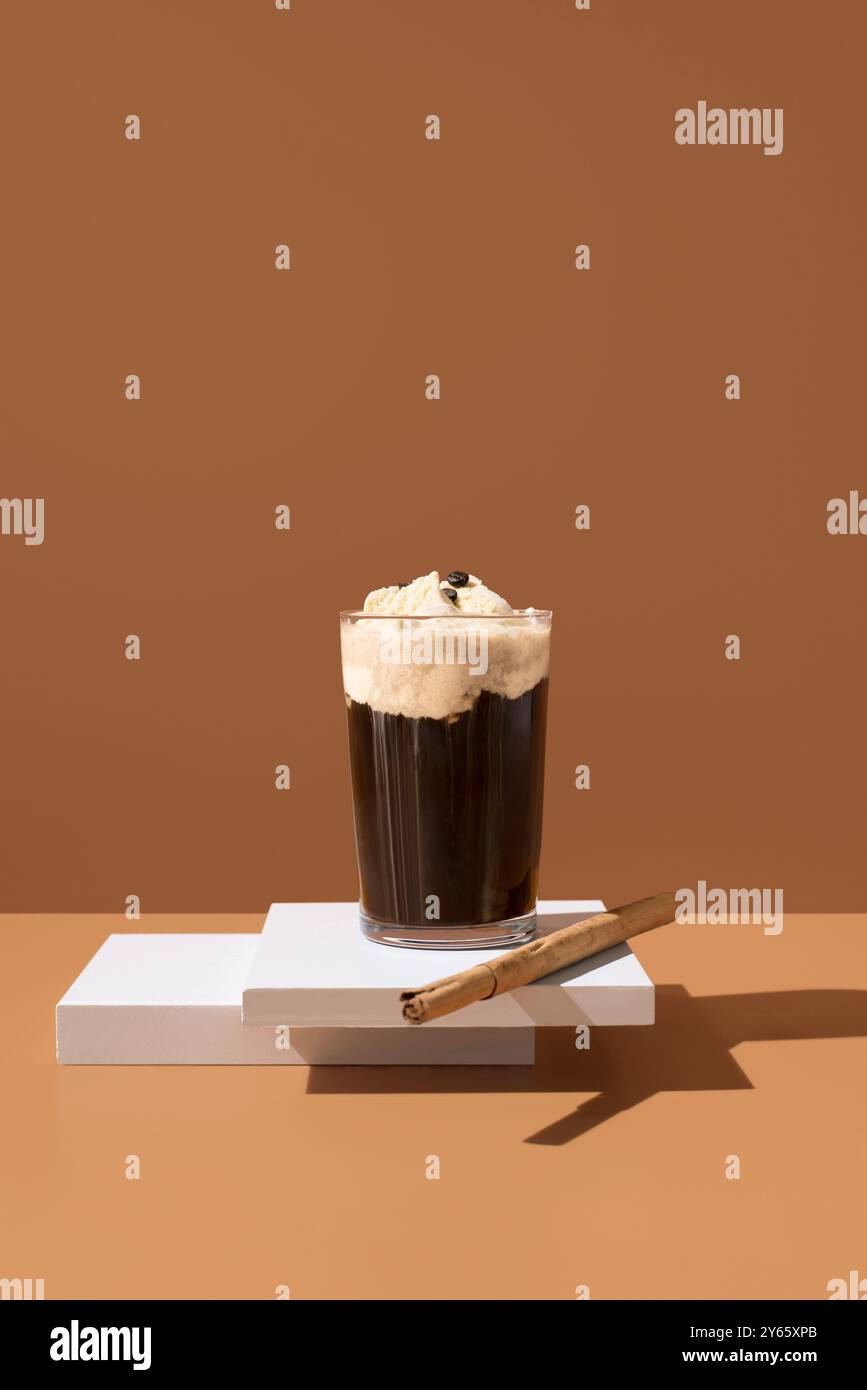 Un delizioso bicchiere di caffè condito con gelato alla vaniglia e uno stecca di cannella, presentato su una piattaforma bianca con un caldo sfondo beige. Foto Stock