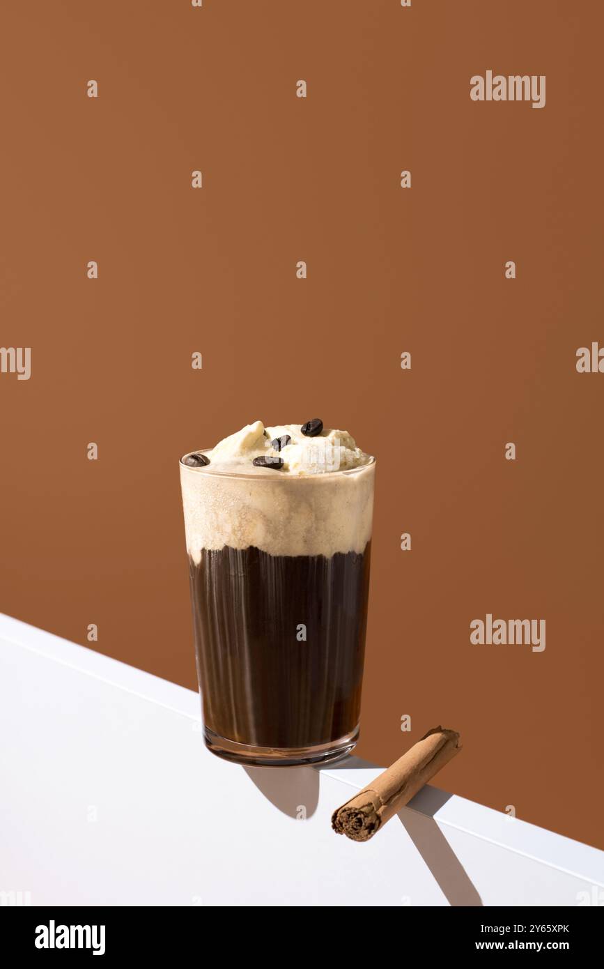 Elegante bicchiere di ricco espresso condito con gelato alla vaniglia e chicchi di caffè, con un bastoncino di cannella sul lato, che evoca un'esperienza di dessert di lusso Foto Stock