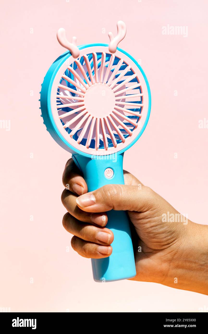 Immagine ravvicinata di una mano che afferra un bizzarro ventilatore portatile blu con stravagante corna rosa su uno sfondo rosa tenue, che illustra un divertimento e un divertimento Foto Stock
