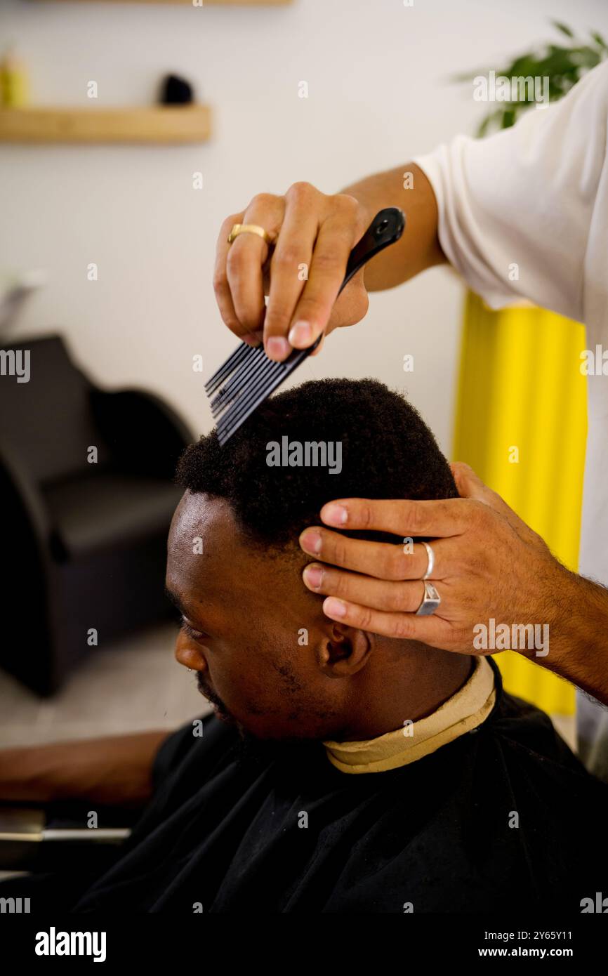 Un barbiere acconcia meticolosamente i capelli di un uomo nero in un ambiente barbiere luminoso e contemporaneo, mettendo in risalto la cura e la cura dei capelli professionali Foto Stock