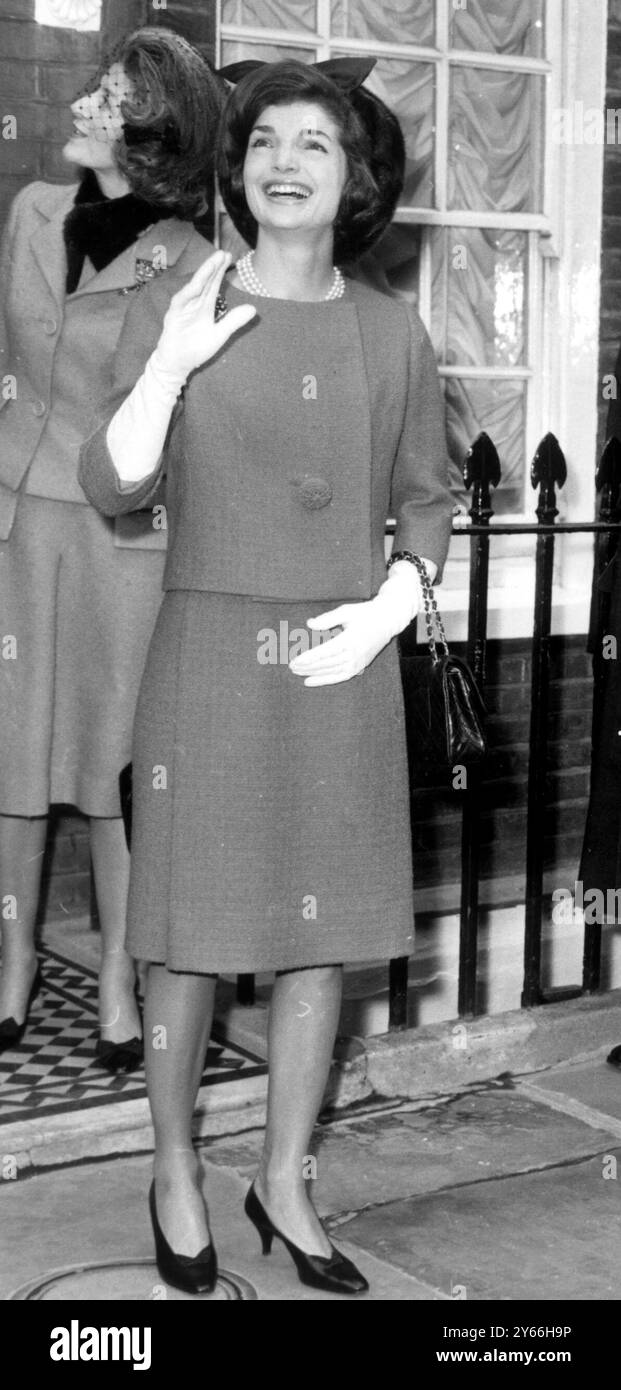 Jackie Kennedy indossò un abito rosa petunia e un cappello in visone, sorridendo e onde mentre guardava un gruppo di operai bilanciati su un'impalcatura da un edificio in costruzione nelle vicinanze mentre lasciava la casa di sua sorella la principessa Lee Radziwill (sfondo). 28 marzo 1962. Foto Stock