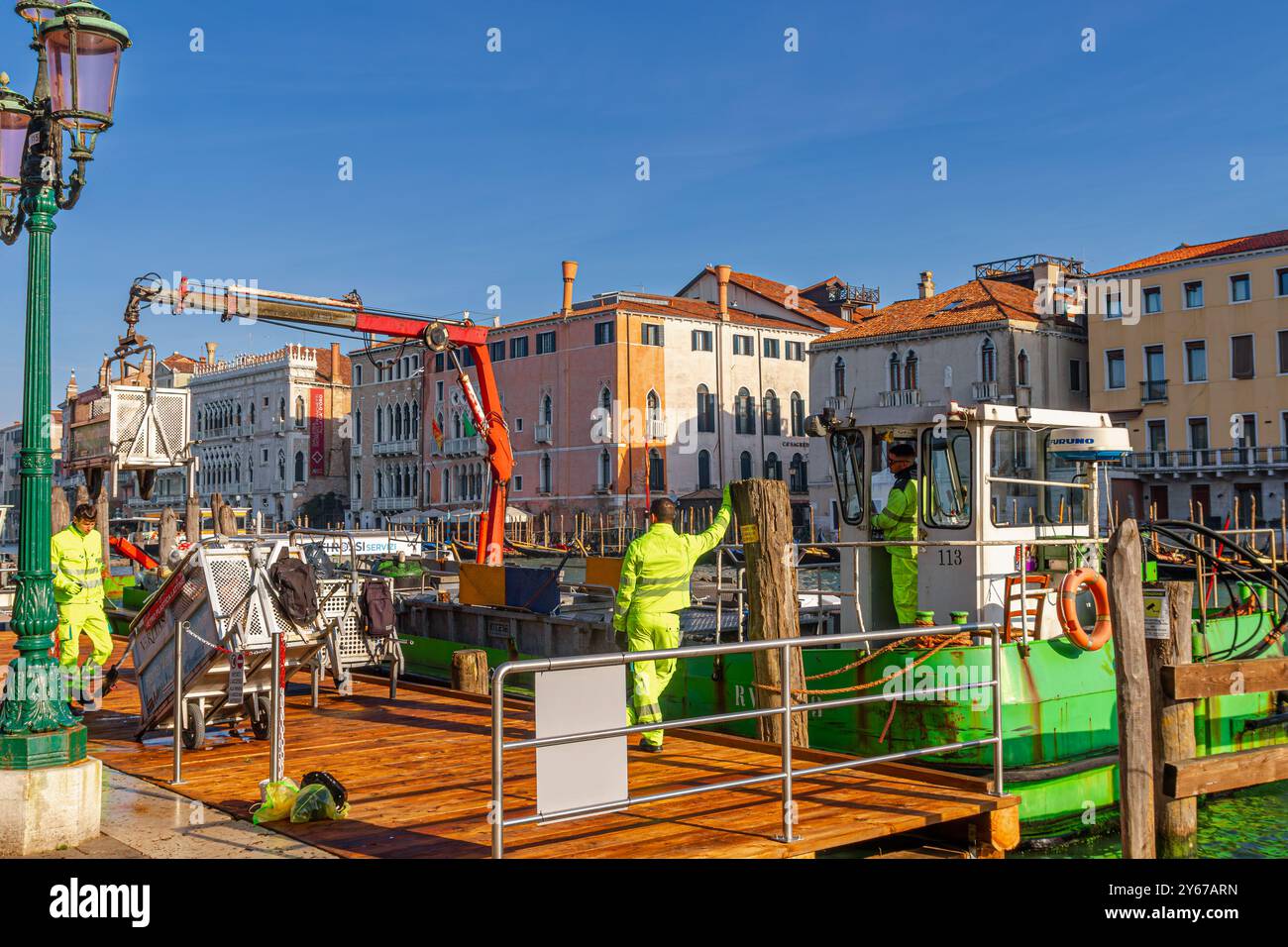 Gli uomini su una barca per rifiuti con braccio di sollevamento idraulico che raccoglie e svuota i contenitori dei rifiuti vicino al mercato di Rialto lungo il Canal grande a Venezia Foto Stock