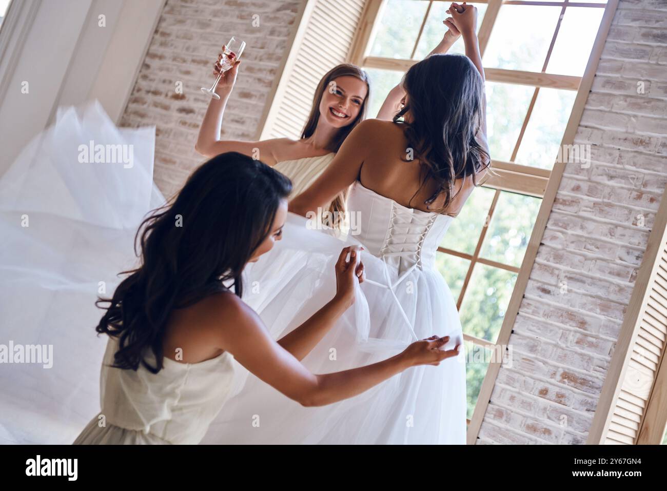 Felice anticipazione. Damigella d'onore che lega un corsetto di un abito da sposa mentre aiuta una bella sposa nel camerino Foto Stock