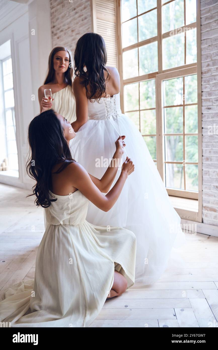 Gli amici di Beat sono sempre in giro. L'intera lunghezza della damigella d'onore che lega un corsetto di un abito da sposa mentre aiuta una bella sposa nel camerino Foto Stock