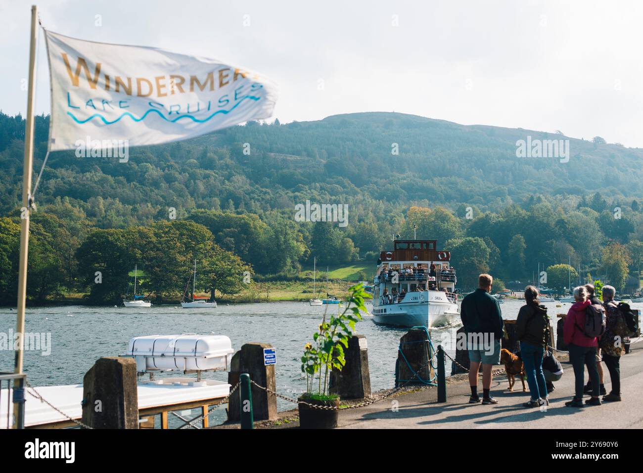 I turisti attendono al molo una barca per le crociere sul lago Windermere nel Lake District in una giornata ventosa, circondato da colline, alberi e acque tranquille. Foto Stock