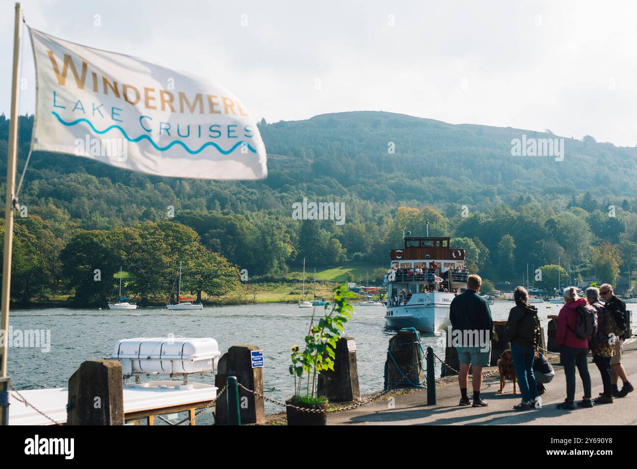 I turisti attendono al molo una barca per le crociere sul lago Windermere nel Lake District in una giornata ventosa, circondato da colline, alberi e acque tranquille. Foto Stock