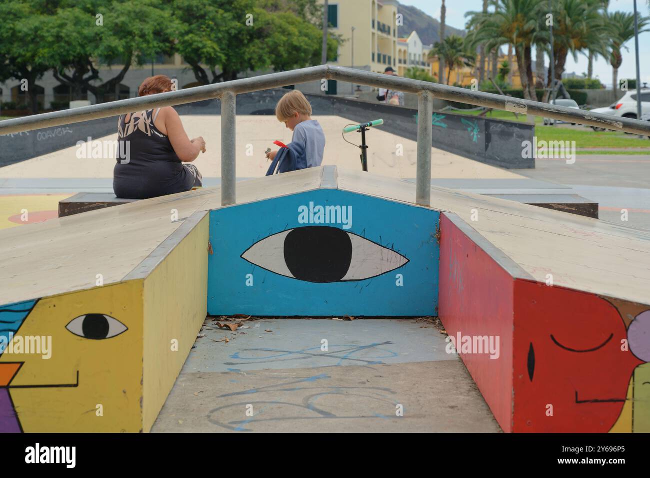 Rampe colorate con volti audaci in uno skatepark di funchal, mentre un ragazzo e una donna si siedono nelle vicinanze Foto Stock