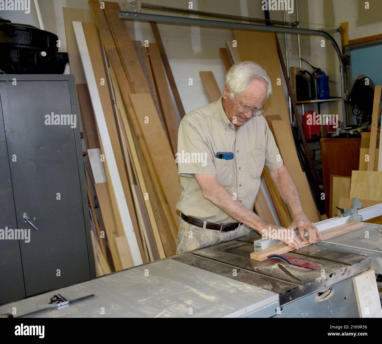 Il maschio senior taglia il rivestimento in legno su una sega da tavolo. È nella sua officina. Indossa pantaloni camo e camicia marrone chiaro. Foto Stock