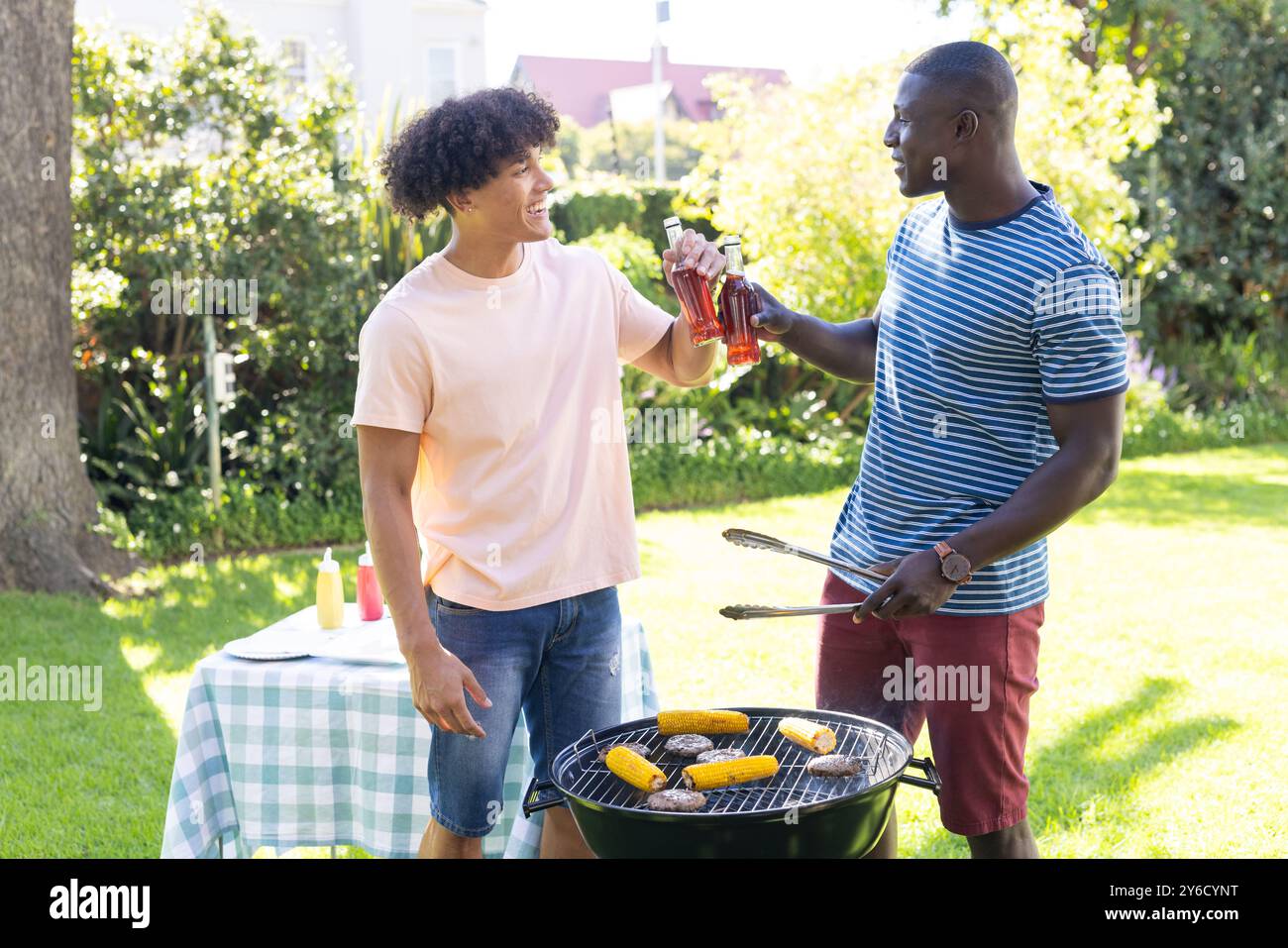 Grigliate e tostatura con bevande, due amici maschi diversi che si godono il barbecue all'aperto Foto Stock