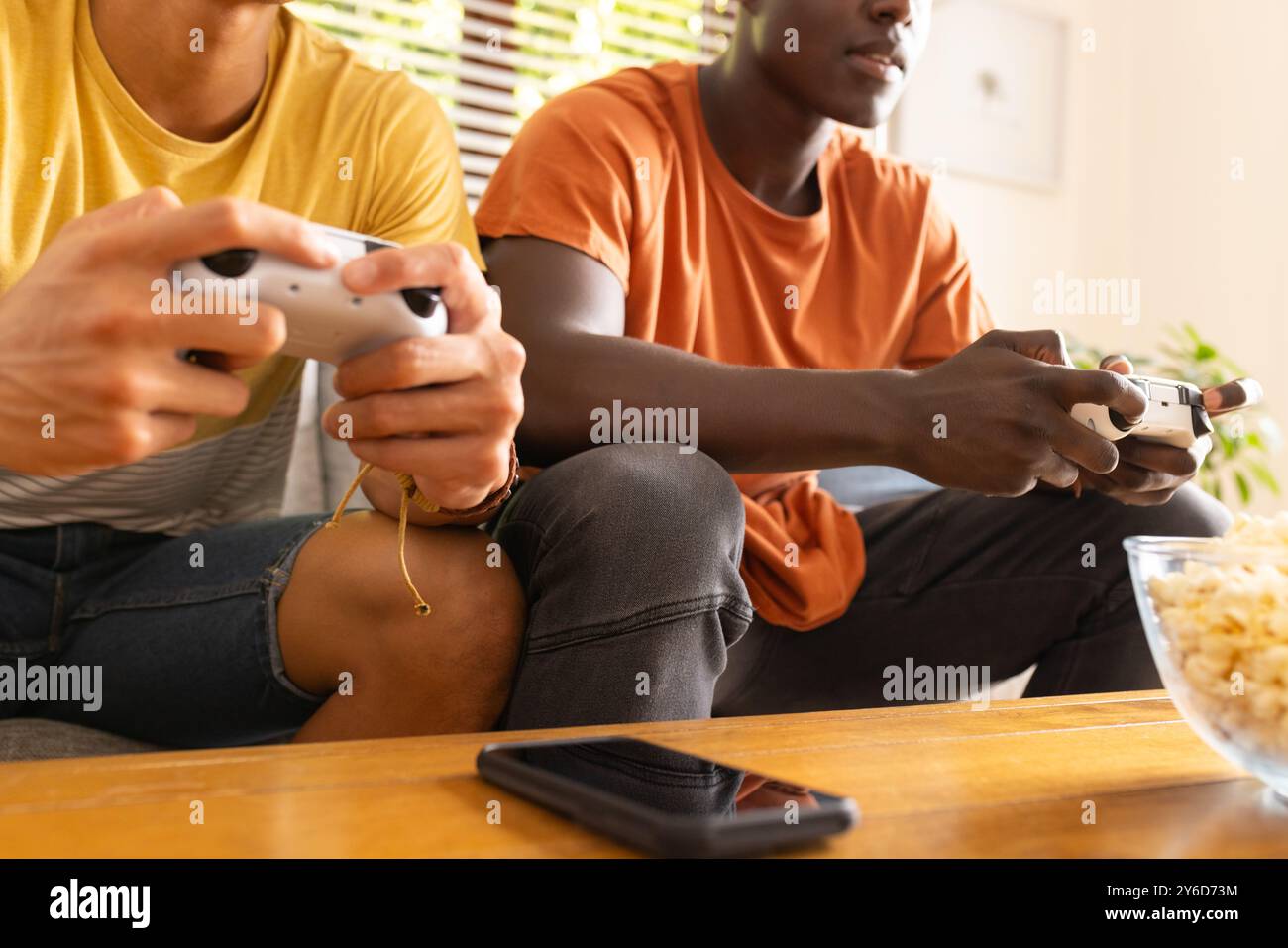 A casa, giocando ai videogiochi, due diversi amici maschi che usano i controller e si siedono sul divano Foto Stock