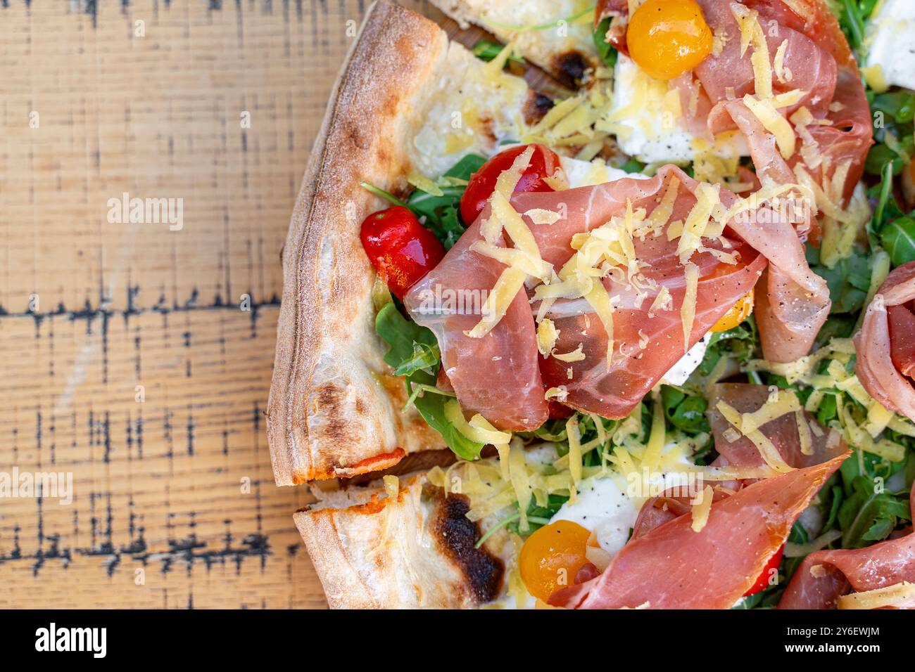 Primo piano di pizza artigianale condita con prosciutto, rucola fresca, pomodori ciliegini gialli e rossi colorati e formaggio grattugiato. Servito su legno rustico Foto Stock