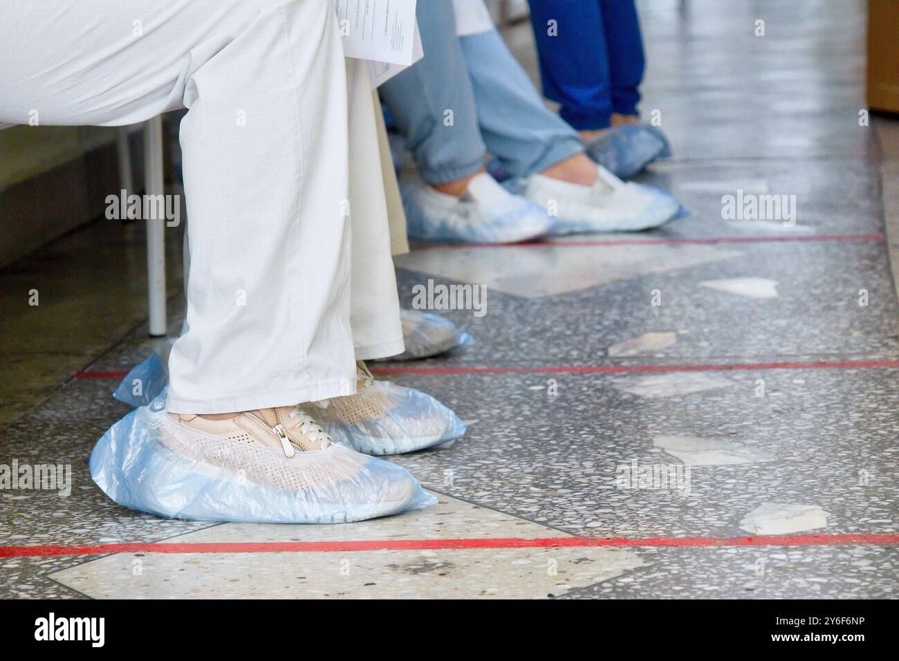 Diverse persone che indossano copriscarpe per uso medico si trovano in fila su un pavimento piastrellato marcato, indicativo di un ambiente sanitario in cui la pulizia e l'ordine sono meticolosamente mantenuti. Foto Stock