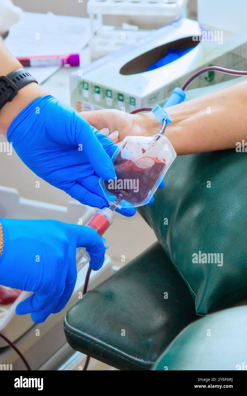 Un operatore sanitario con guanti blu raccoglie sapientemente una donazione di sangue da un paziente seduto comodamente. L'ambiente è una struttura medica attrezzata per la donazione di sangue, che sottolinea l'importanza della salute della comunità. Foto Stock