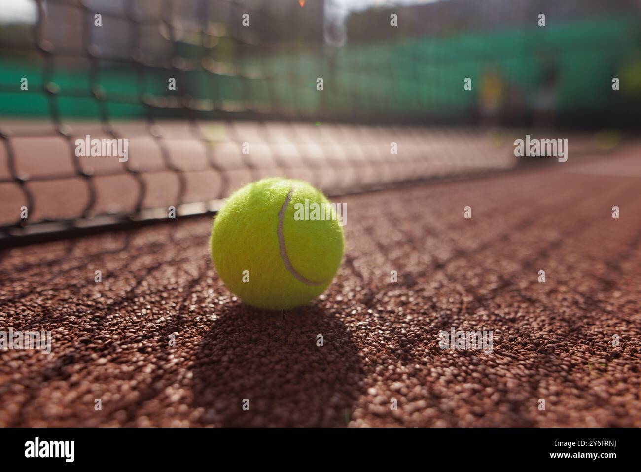 Primo piano di una palla da tennis appoggiata su un campo di argilla, con una rete in primo piano e giocatori sfocati sullo sfondo Foto Stock