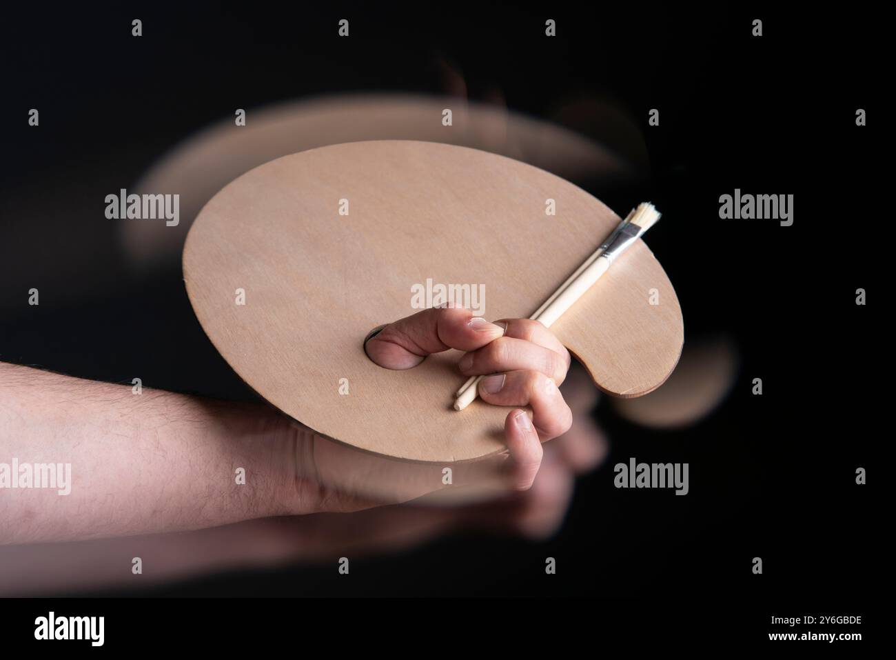 Primo piano della mano dell'uomo che tiene la tavolozza rotonda di legno e pennelli con effetto di movimento su sfondo nero Foto Stock