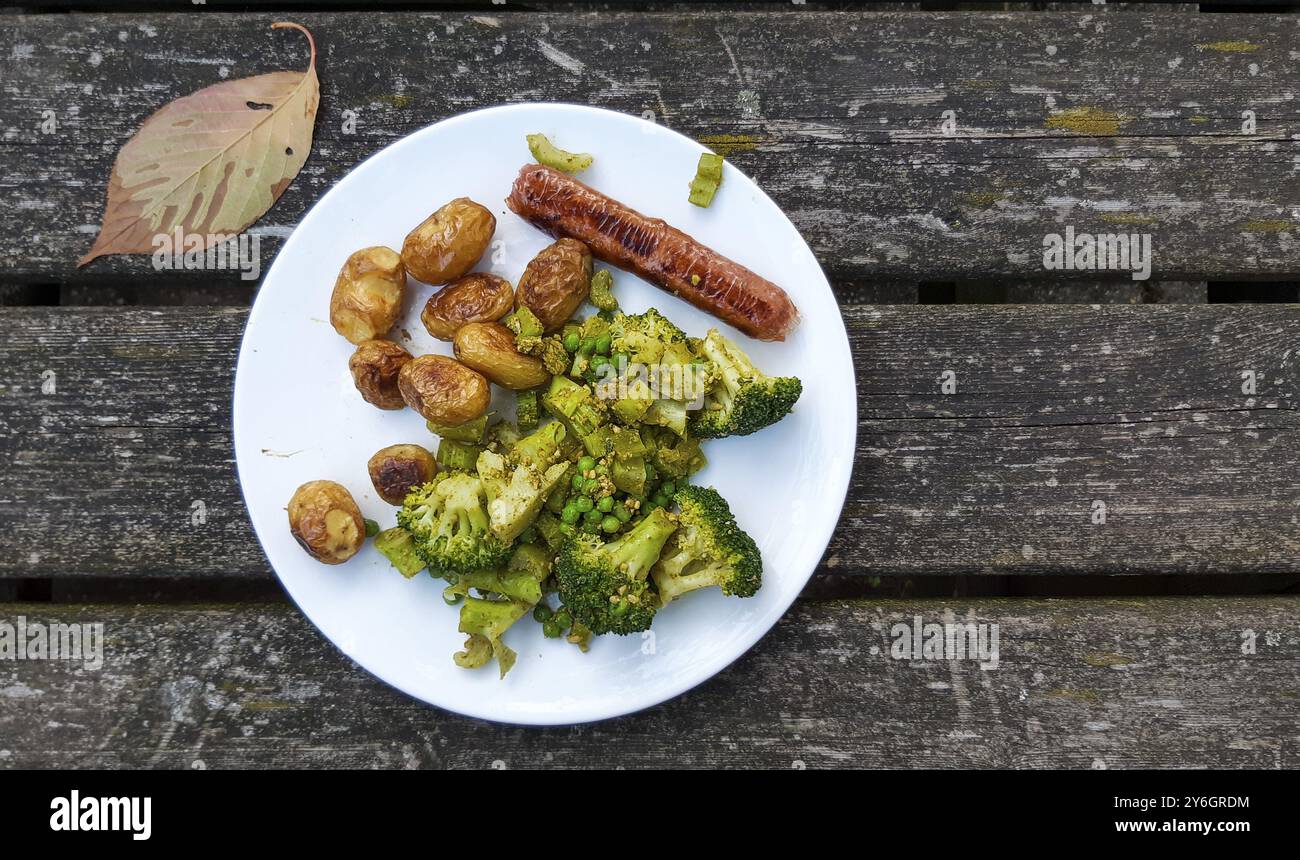 Pasto vegano con potatos al forno, sausace a base di piante, broccoli, piselli e pesto su un piatto bianco. Veganismo e cibo delizioso su legno intemperie ta Foto Stock