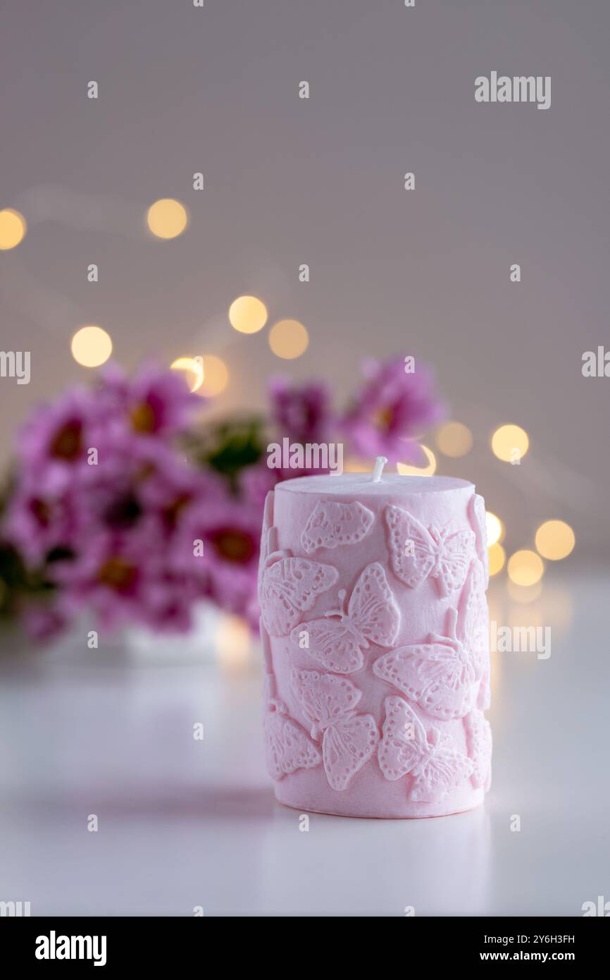 Candela decorativa rosa con motivo a farfalla. Fiori e luci sullo sfondo con spazio di copia. Arredi per la casa. Interni accoglienti. Preparazione di candele di cera di soia. Foto Stock
