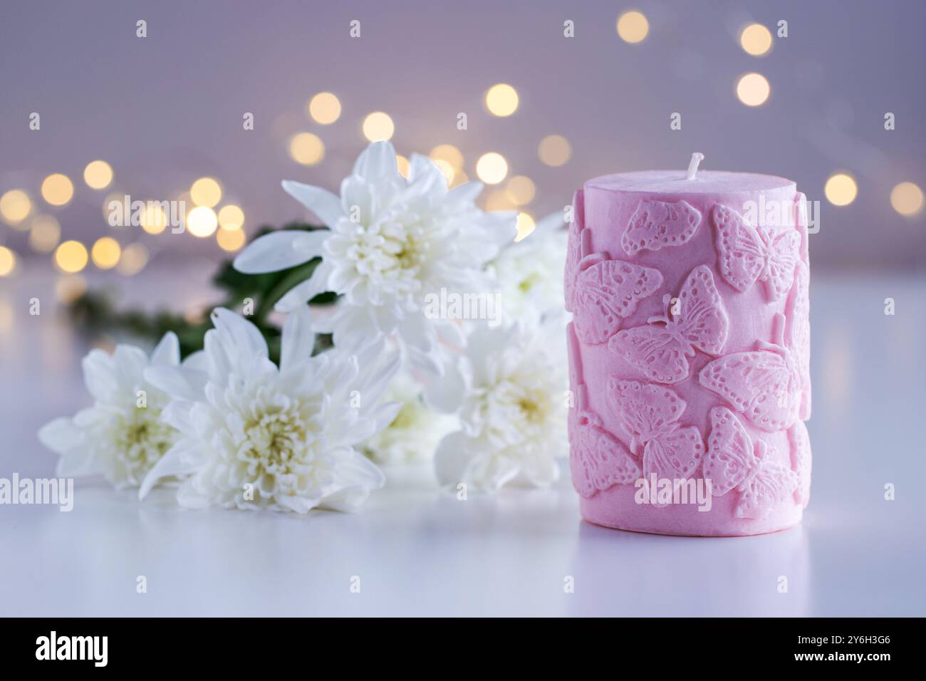 Candela decorativa rosa con motivo a farfalla. Fiori e luci sullo sfondo con spazio di copia. Arredi per la casa. Interni accoglienti. Preparazione di candele di cera di soia. Foto Stock