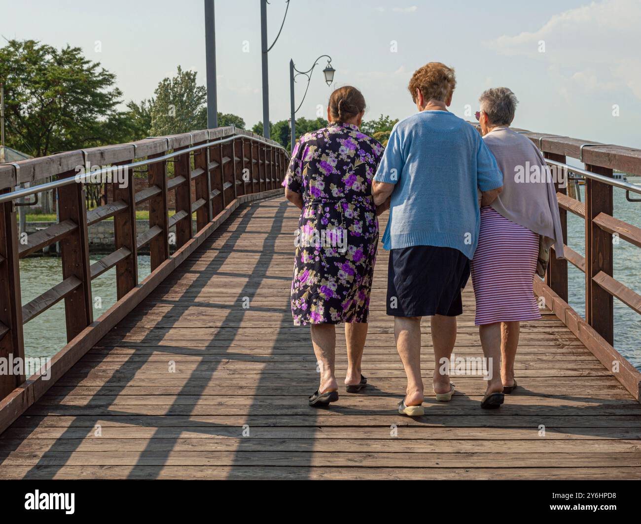 Tre donne bianche più anziane con i capelli grigi che camminano attraverso un ponte di legno con le braccia collegate. Foto Stock