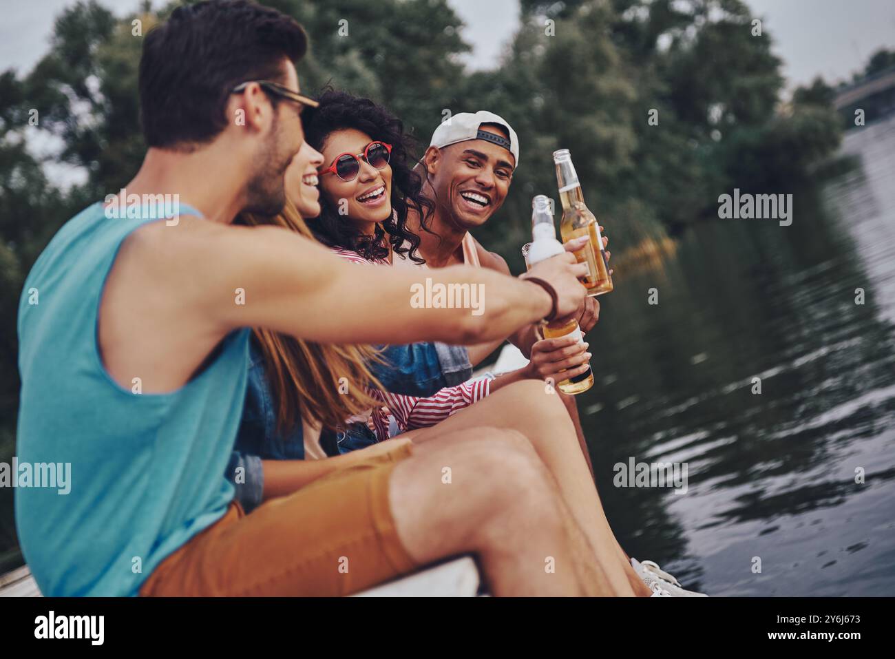 Ci stiamo divertendo insieme. Gruppo di giovani felici in abbigliamento casual sorridenti e bevono birra mentre si siede sul molo Foto Stock