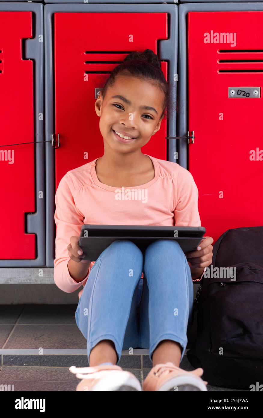 Seduta accanto agli armadietti, ragazza che usa il tablet e sorride nel corridoio della scuola Foto Stock