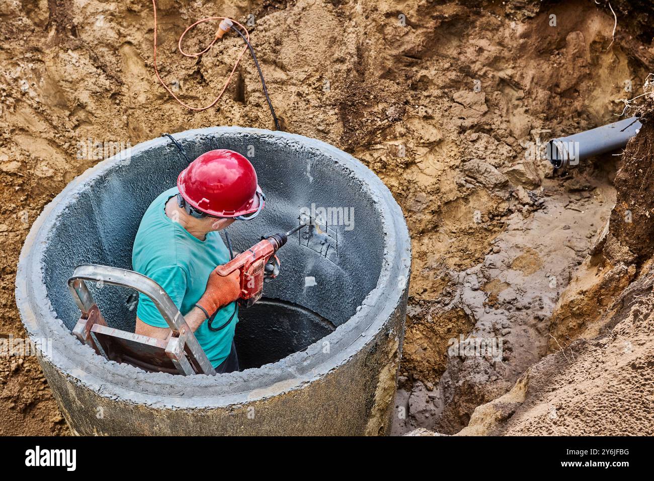 Il lavoratore utilizza un martello perforatore con cordoncino con scalpello per punzonare l'anello in calcestruzzo del serbatoio settico. Foto Stock