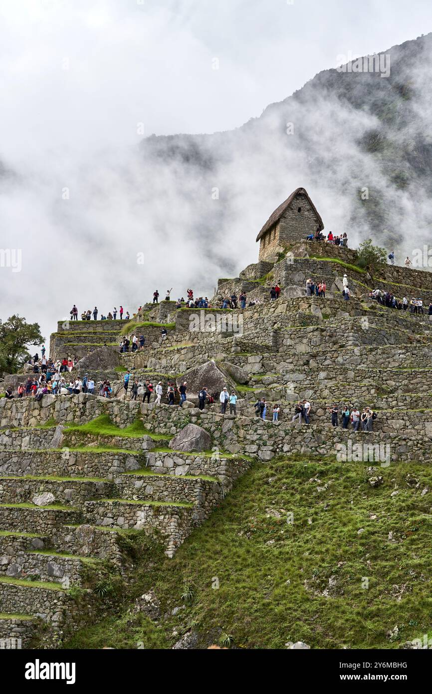 Una vista panoramica di Machu Picchu con turisti che esplorano le iconiche rovine inca, circondati da nebbia e lussureggianti montagne verdi. Foto Stock