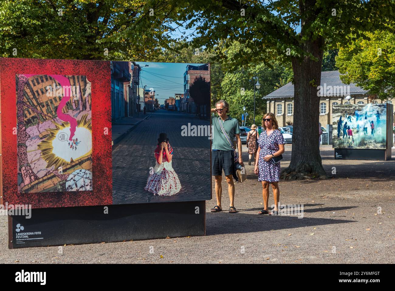 Un uomo e una donna passano davanti a un display a due pannelli al Landskrona Photo Festival 2024 che mostra immagini della serie “Shine Heroes” dell'artista uruguayano Federico Estol: un'esplosione disegnata a mano in stile fumetto accanto a una fotografia di una figura inginocchiata e ammantata su una strada acciottolata, e un ragazzo in piedi da solo su una strada deserta a La Paz o El Alto, in Bolivia. Estol ha lavorato con sessanta ragazzi lustrascarpe delle due città, che hanno aiutato a mettere in scena le scene come produttori e protagonisti di un saggio fotografico mirato a combattere lo stigma sociale legato al loro mestiere. Citadellstaden, Skåne, Svezia. Foto Stock