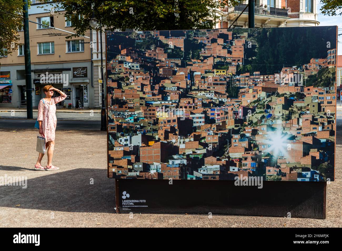 Una passante si protegge gli occhi mentre passa davanti a una grande fotografia della serie "Shine Heroes" dell'artista uruguayano Federico Estol al Landskrona Photo Festival 2024, che mostra un quartiere collinare densamente costruito di La Paz o El Alto, in Bolivia. Estol ha lavorato con sessanta ragazzi lustrascarpe delle due città, che hanno aiutato a mettere in scena le scene come produttori e protagonisti di un saggio fotografico mirato a combattere lo stigma sociale legato al loro mestiere. Citadellstaden, Skåne, Svezia. Foto Stock