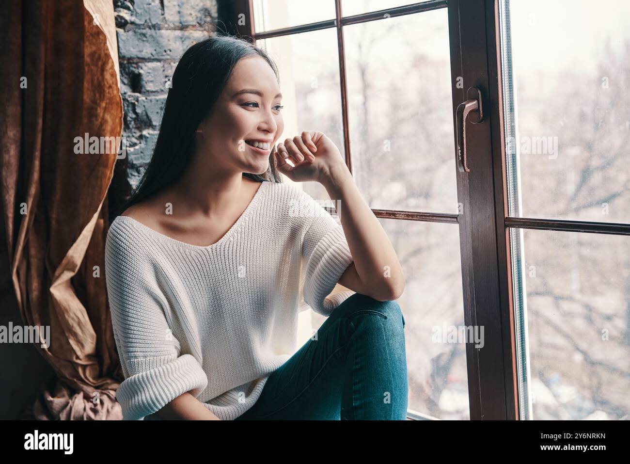 Bei ricordi. Una giovane donna attraente sorridente e che guarda lontano mentre si siede sul davanzale della finestra a casa Foto Stock
