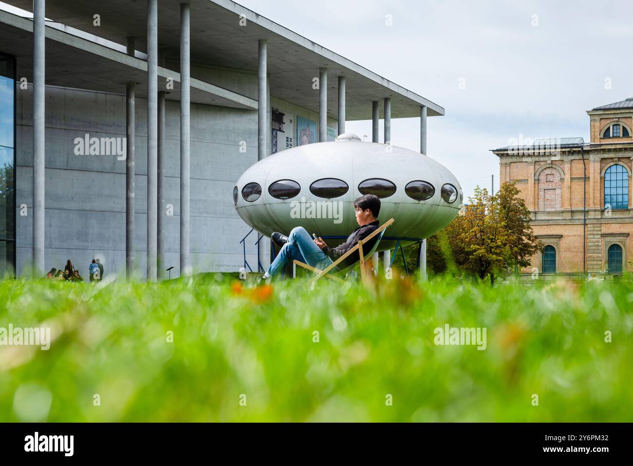 Un giovane uomo siede su una sdraio sul prato di fronte alla Pinakothek der moderne e all'installazione artistica futuro House di Matti Suuronen, Monaco di Baviera Foto Stock