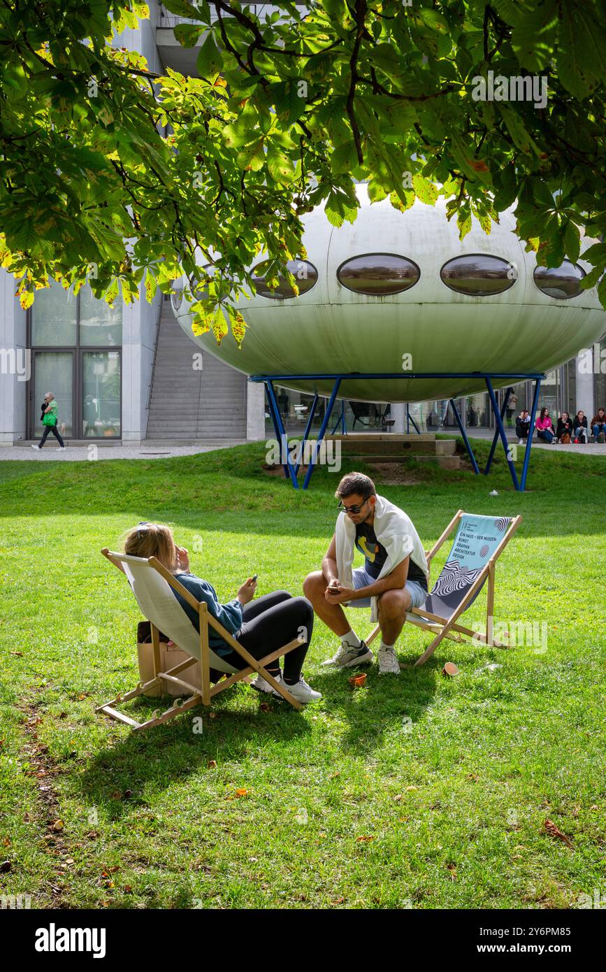 Una coppia siede su sdraio sul prato di fronte alla Pinakothek der moderne e all'installazione artistica futuro House di Matti Suuronen, Monaco di Baviera Foto Stock