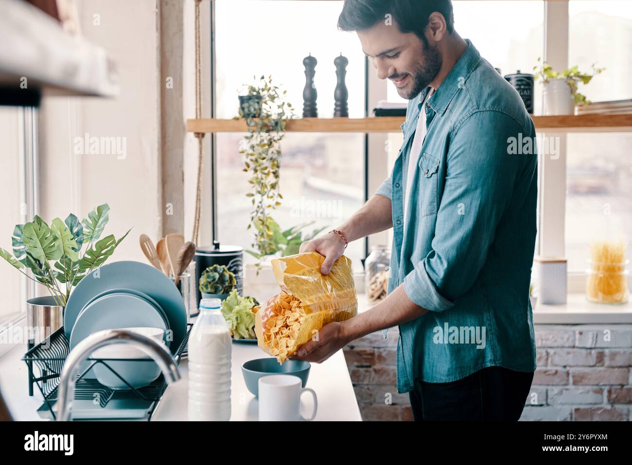Colazione facile. Bel giovane uomo in abbigliamento casual che prepara fiocchi di mais e sorride mentre sta in cucina a casa Foto Stock