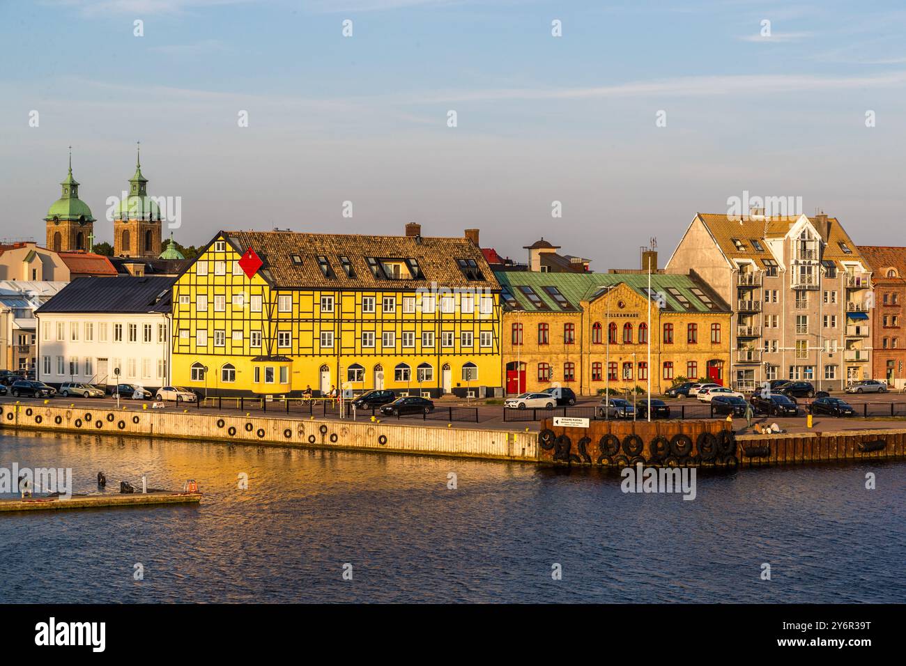 Un edificio in legno a graticcio giallo con una persiana rossa domina il lungomare di Landskrona, con le guglie verdi di una chiesa che si ergono dietro di esso. Auto sono parcheggiate lungo il molo storico. Skåne, Svezia. Foto Stock
