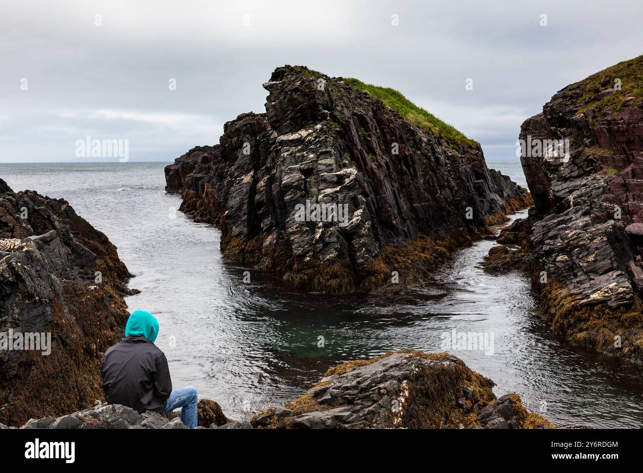 Ragazzo seduto vicino all'oceano guardando il mare e le rocce in una giornata fresca e nuvolosa a Terranova Foto Stock