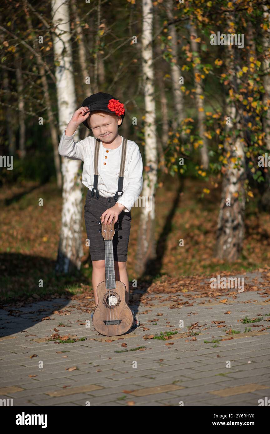 Un ragazzo in abito vintage con una piccola chitarra, in piedi su un sentiero lastricato con alberi di betulla sullo sfondo. Foto Stock