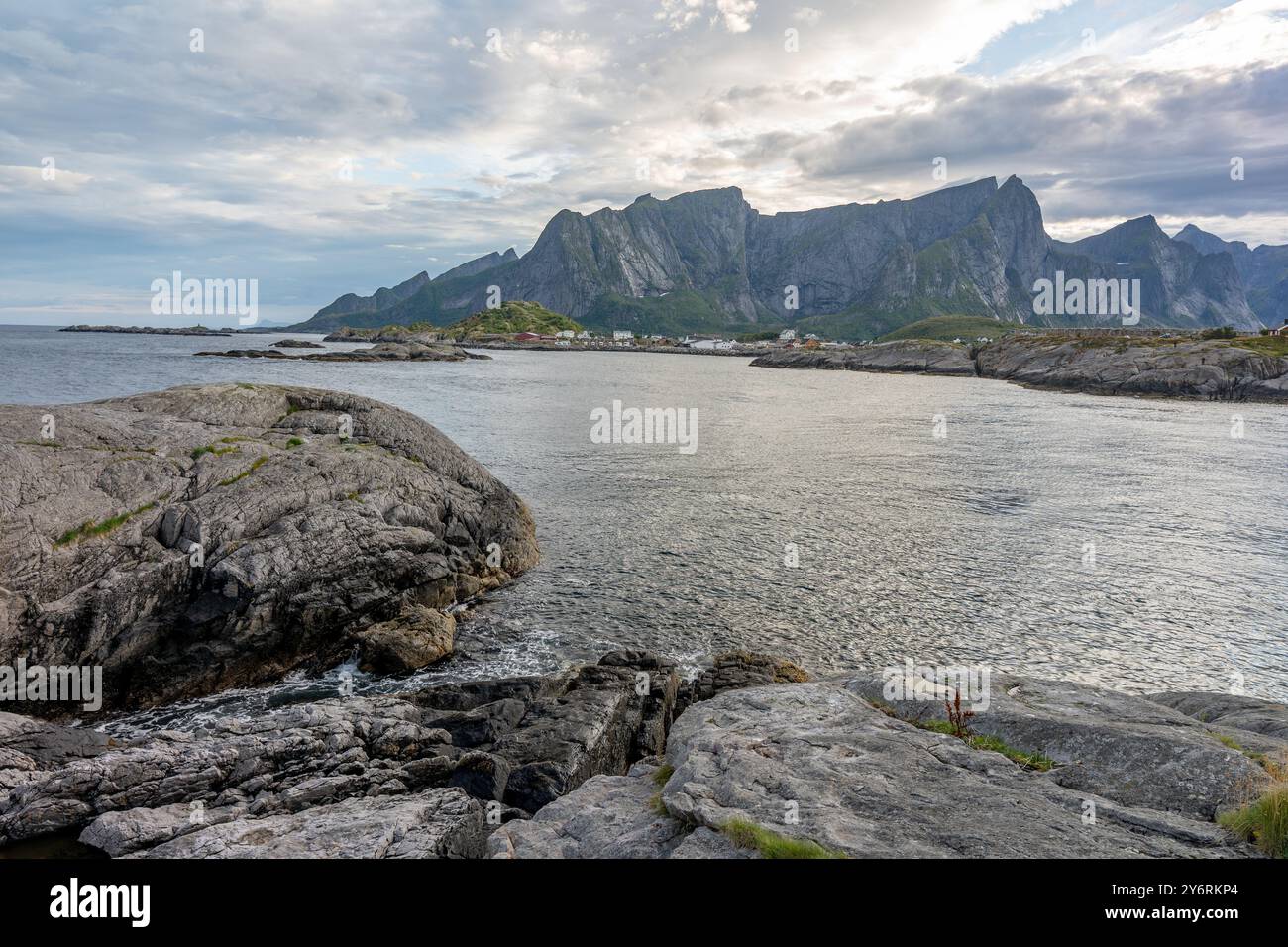 Piccola città vicino al lago vista dalla spiaggia rocciosa opposte e montagne rocciose sullo sfondo, Lofoten Norvegia Foto Stock