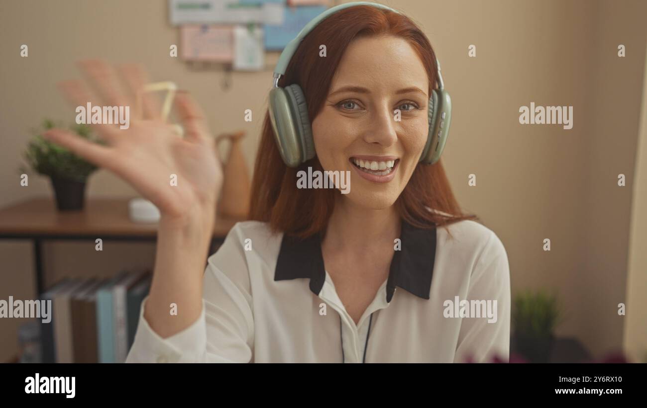 Una giovane donna sorridente con le cuffie gode di una videochiamata nel suo accogliente ufficio domestico. Foto Stock
