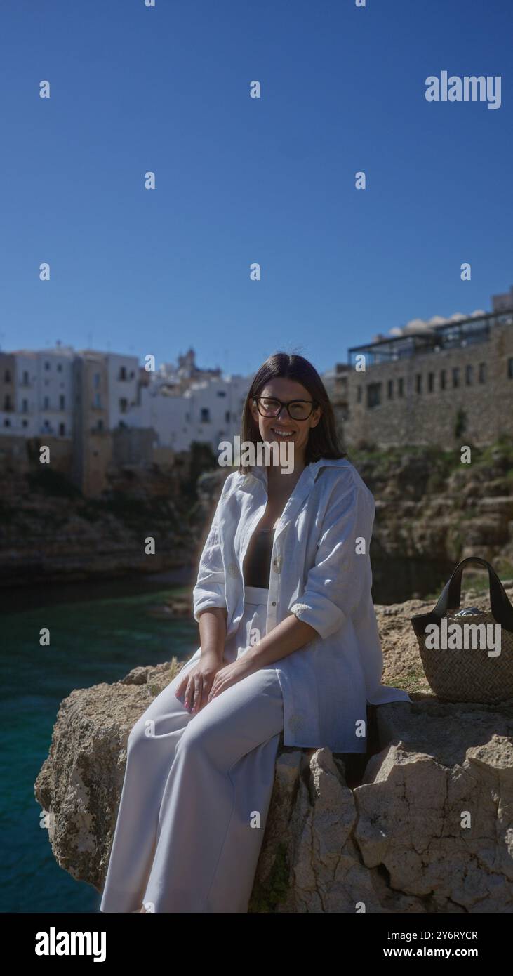 Una giovane donna ispanica siede su una scogliera rocciosa che si affaccia sullo splendido mare, con polignano a mare in puglia, italia sullo sfondo in una giornata di sole. Foto Stock