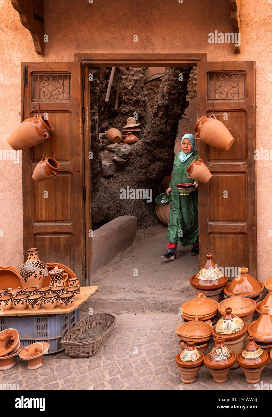 Una donna musulmana che indossa abiti tradizionali e hijab che porta ceramiche si trova all'ingresso di un tradizionale laboratorio di suk marocchino. La porta è ado Foto Stock