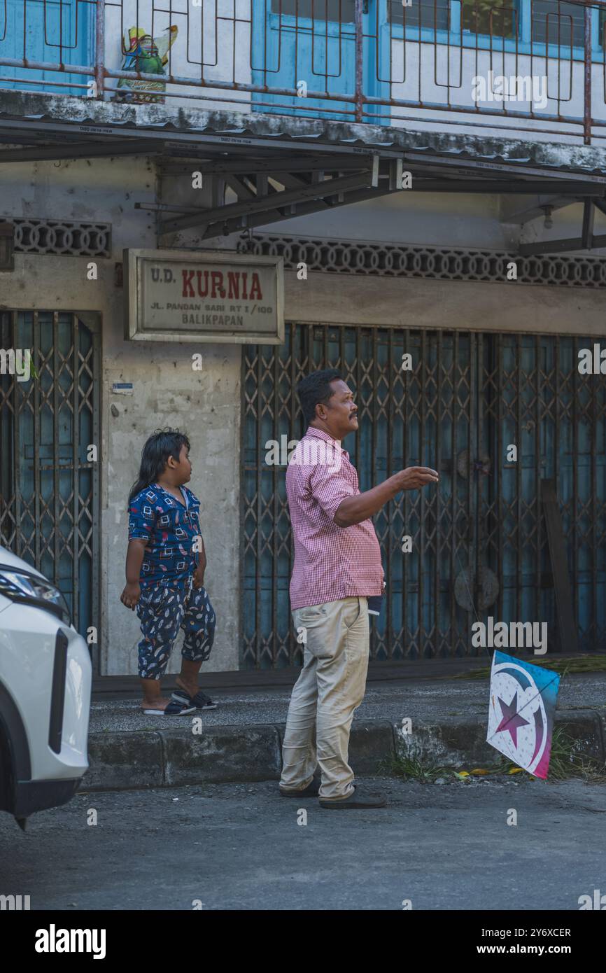 Balikpapan, Indonesia - 26 giugno 2024. Con un aquilone colorato in mano, il padre si trova vicino al marciapiede, suo figlio al suo fianco. Foto Stock
