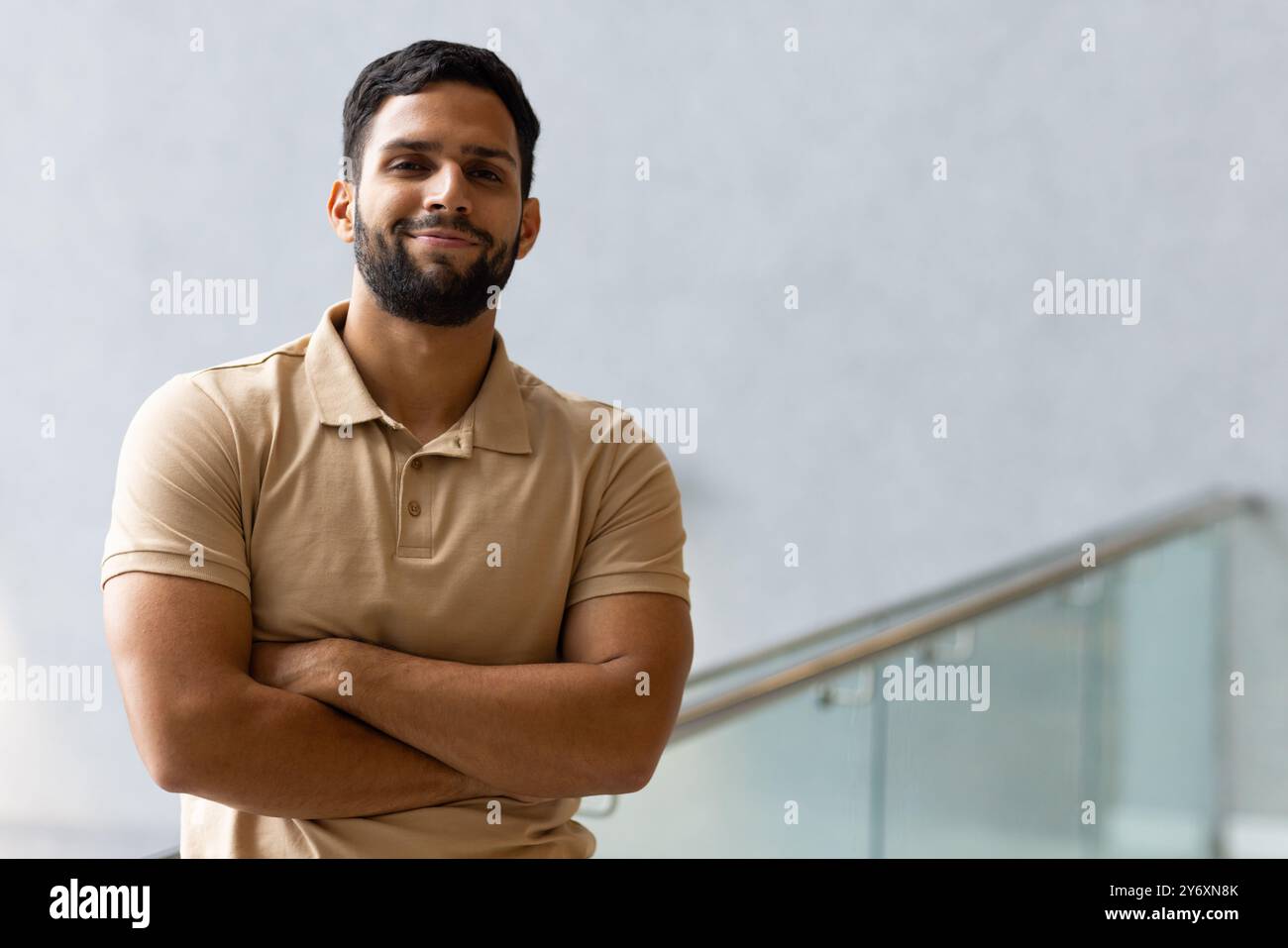 Uomo sicuro di sé che incrocia le braccia e sorride, in piedi all'interno vicino a ringhiere di vetro, copia spazio Foto Stock