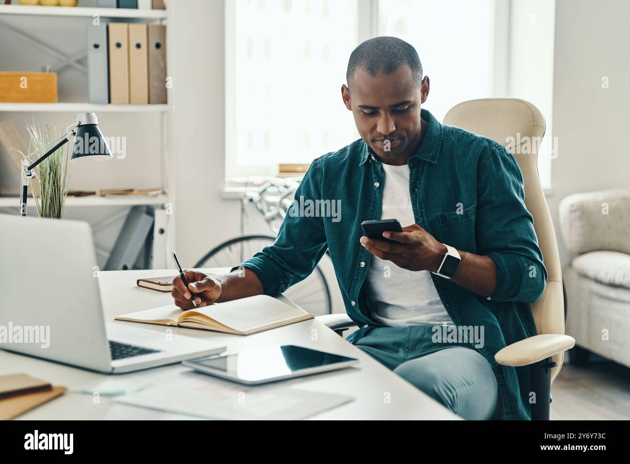 Controllo dei messaggi. Giovane uomo africano in camicia che usa lo smartphone mentre è seduto in ufficio Foto Stock