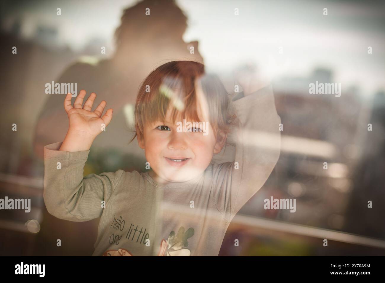 Un bambino gode di un momento giocoso vicino alla finestra, salutando con entusiasmo. La luce del sole si filtra, illuminando l'espressione allegra sulla loro f Foto Stock