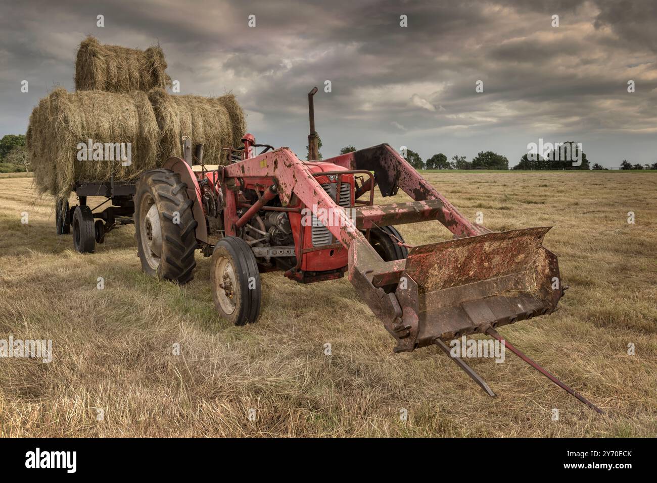 Un trattore d'epoca viene utilizzato per raccogliere balle di fieno, mentre si avvicinano le nuvole di tempesta, nel Wiltshire, in Inghilterra. Foto Stock